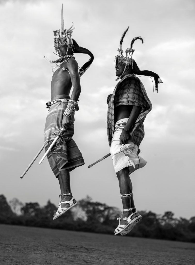 Two men in traditional attire jump mid-air showcasing cultural dance with headdresses in Kenya.