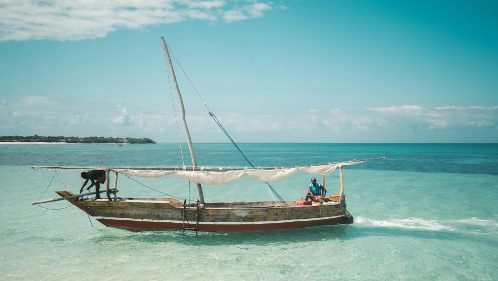 Two local fishermen navigate a traditional dhow on the clear waters of Zanzibar under a bright blue sky.
